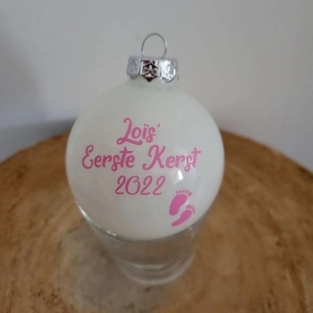 Witte handgemaakte kerstbal met roze tekst Baby's Eerste Kerst hangend in een kerstboom, Gepersonaliseerde witte kerstbal voor baby's eerste kerst in een sfeervolle kerstsetting, Baby's eerste kerstbal met naam en jaartal in blauw of roze tekst